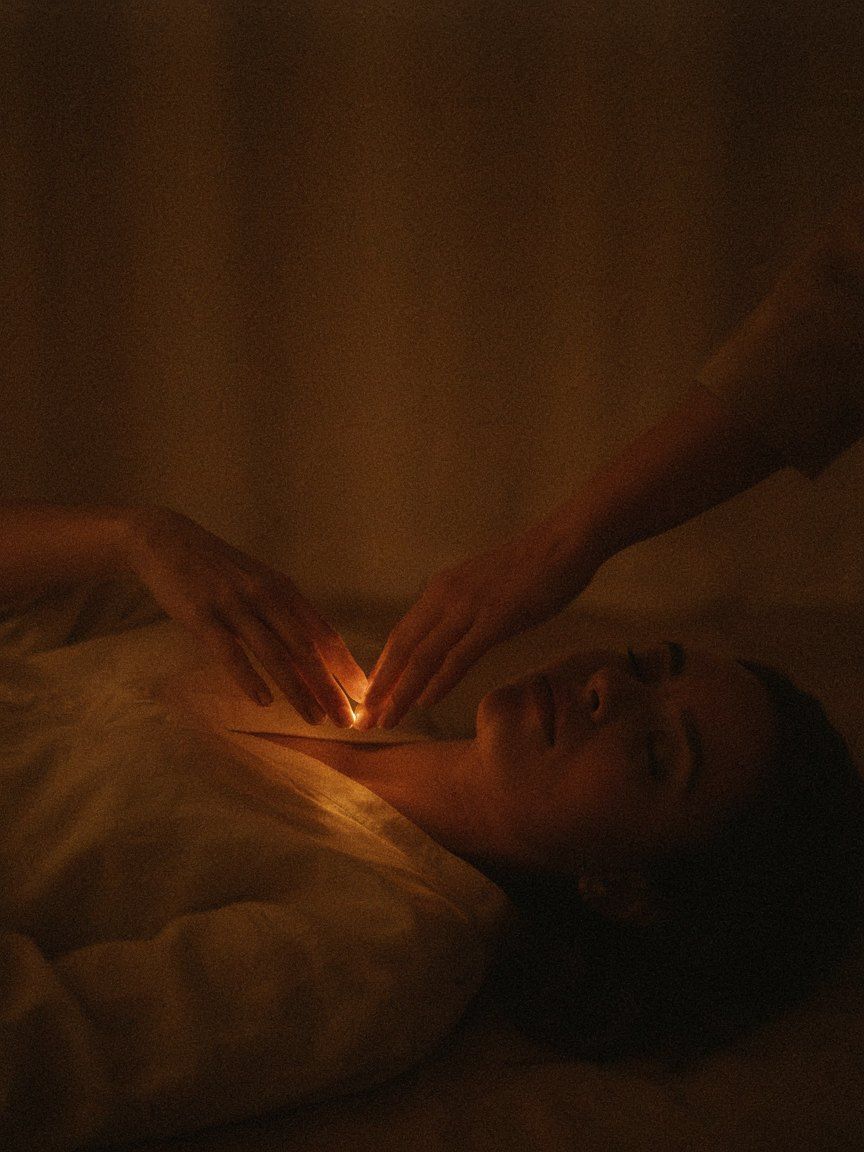 Two hands hovering just above a resting woman&rsquo;s chest, warm candlelight pooling in the space between